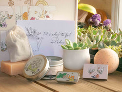 It's okay feel not okay Succulent Gift Box with natural soap, bath bomb, candle, custom matches, lavender sachet and lip balm, in front of a decorative gift box