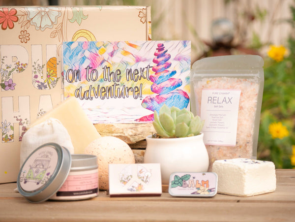 On to the next adventure Succulent Gift Box with relax bath salt, natural soap, bath bomb, shower steamer, candle, custom matches, lavender sachet and lip balm, in front of a decorative gift box